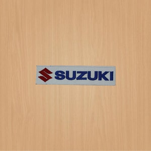 May include: A white embroidered patch with the word "SUZUKI" in blue lettering and a red "S" logo.
