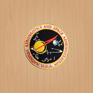 Puede incluir: Un parche bordado en blanco y negro con el texto "National Aeronautics and Space Administration" y "U.S.A." que rodea un cohete rojo y blanco y un planeta amarillo con un anillo a su alrededor.