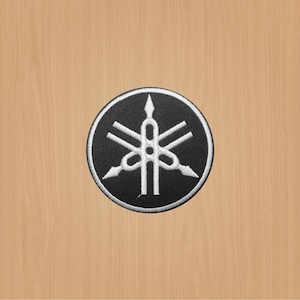 Könnte beinhalten: Ein gestickter schwarzer und weißer Aufnäher mit dem Yamaha-Logo. Das Logo ist eine stilisierte Stimmgabel mit drei Zinken auf jeder Seite.