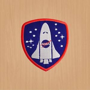 May include: A blue and red embroidered patch featuring a white space shuttle design. The shuttle has the NASA logo on the side and is surrounded by white stars on a blue background.
