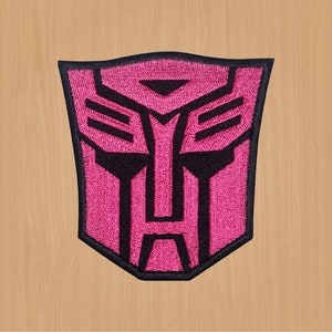 May include: Pink and black embroidered iron-on patch featuring the Autobot symbol from the Transformers franchise.