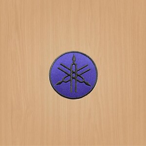 May include: A purple embroidered patch featuring the Yamaha tuning fork logo. The logo is black and depicts three tuning forks, with a central tuning fork that is longer than the other two.