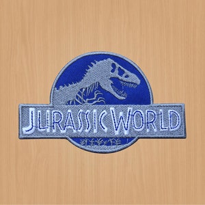 Może przedstawiać: Naszywka haftowana w kolorze niebieskim i szarym z logo Jurassic World. Logo przedstawia sylwetkę dinozaura i napis "Jurassic World".