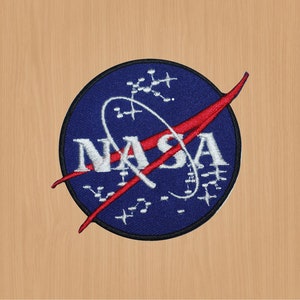 May include: A blue and red embroidered patch featuring the NASA logo. The logo shows a white stylised letter 'N' with a red curved line through it, followed by the letters 'ASA' in white. The patch is encircled by a white circle with a pattern of small white dots and lines.