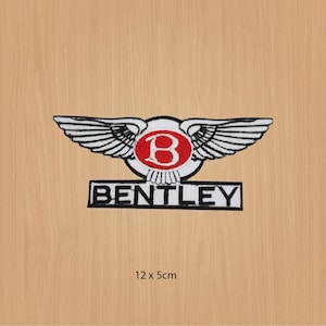 Puede incluir: Un parche bordado blanco y negro con un círculo rojo que contiene una letra "B" blanca y la palabra "BENTLEY" en letras negras. El parche presenta un par de alas con plumas blancas y contornos negros. El parche mide 12 cm por 5 cm.