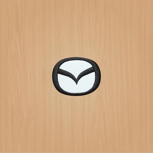 Pode incluir: Um remendo bordado com o logotipo da Mazda. O logotipo é uma forma de asa estilizada dentro de um círculo.
