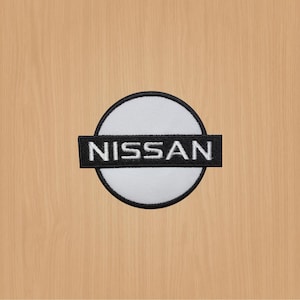 Peut inclure: Un cercle blanc avec une bordure noire et le mot "NISSAN" en lettres blanches sur fond noir.