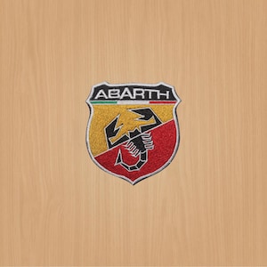 Könnte beinhalten: Ein gestickter schwarzer und weißer Aufnäher mit dem Wort "ABARTH" in weißen Buchstaben auf einem schildförmigen Hintergrund. Der Schild ist in zwei Abschnitte unterteilt, einen gelben und einen roten, mit einem schwarzen Skorpion in der Mitte. Die italienische Flagge ist oben auf dem Schild sichtbar.