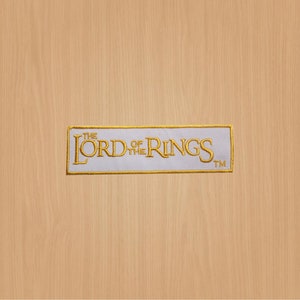 Könnte beinhalten: Weißer gestickter Aufnäher mit goldener Schrift, die "The Lord of the Rings" mit einem Warenzeichen-Symbol lautet.