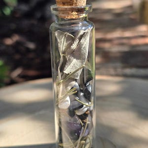 May include: A small glass bottle with a cork stopper filled with white sage, black and white stones, and amethyst crystals.