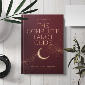 May include: A burgundy book with gold lettering titled "The Complete Tarot Guide" by M.F. Winters. The book cover features a crescent moon and gold glitter.