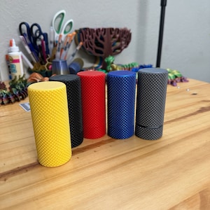 May include: A collection of six cylindrical objects in various colors: yellow, black, red, blue, and gray. Each object has a textured, diamond-patterned surface. They are arranged on a wooden surface, with craft supplies visible in the background.