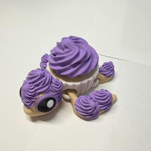 May include: A whimsical, cupcake-shaped figurine with a light tan base, white cup, and a large swirl of lavender frosting. The figurine has two large black and white eyes and lavender frosting accents.
