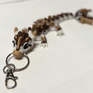May include: A 3D-printed keychain featuring a stylized lizard in white and brown. The lizard has a detailed, segmented body and is attached to a silver keyring and clasp. The keychain is designed to be a decorative accessory.