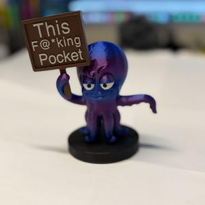 May include: A small, purple and blue gradient octopus figurine standing on a black circular base. The octopus holds a brown rectangular sign with white text that reads, "This F@*king Pocket," and features a cartoonish expression with large eyes.