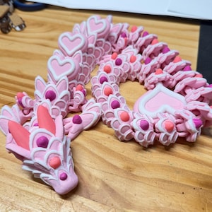 May include: A pink and white articulated dragon figurine with heart-shaped details. The dragon has a detailed design with connected segments, each with heart and circular details. The dragon's head is prominent, showcasing intricate features. The figurine is on a wooden surface.