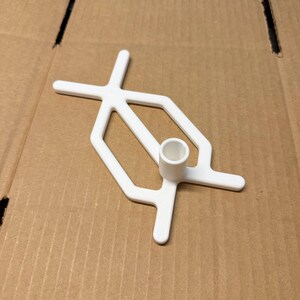May include: A white plastic object with a cross-shaped top and a cylindrical base. The object has a geometric design and is set against a brown cardboard background. The object appears to be a tool or a component.