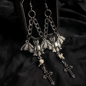 May include: A pair of silver-toned drop earrings featuring a bat charm suspended from a metal chain. Below each bat is a small, off-white bone-like segment connected to a silver-toned cross pendant. The earrings hang from standard metal hooks.