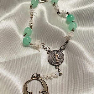 May include: A rosary with light green beads and small white bone-shaped accents. The rosary features a metal pendant with a bat design and a circular medallion with a religious figure. The rosary is displayed on a white satin fabric.