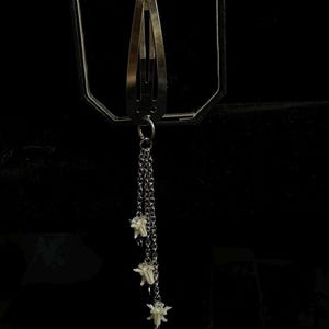 May include: A silver-toned hair clip with three dangling chains, each ending in a small, white, flower-like charm. The clip is a classic barrette style, set against a dark background.