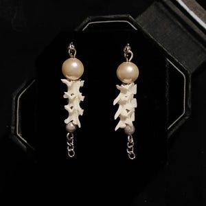 以下が含まれることがあります： 真珠がついた白い骨のような形のイヤリングのペア。イヤリングの下には銀色のチェーンが付いている