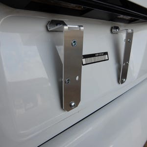 以下が含まれることがあります： 白い車のナンバープレートブラケット2つのクローズアップ。ブラケットは車のバンパーに取り付けられており、シルバー仕上げです。ブラケットはナンバープレートを保持するように設計されています。
