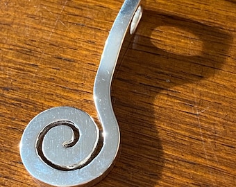 May include: A silver pendant with a spiral design. The pendant has a curved, elongated top with a small loop for a chain. The spiral is centered and has a smooth, polished finish. The pendant is likely made of sterling silver.