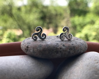May include: A pair of silver stud earrings featuring a Celtic knot design. The earrings are small and delicate, and they are set against a background of rocks.