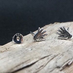 Op de afbeelding: Een paar zilveren oorbel studs met een handgebaar ontwerp. De oorbellen worden getoond op een stuk drijfhout.