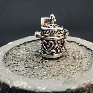 May include: A silver charm shaped like a small, ornate box with a hinged lid. The box is decorated with intricate designs and a rope-like border around the top.