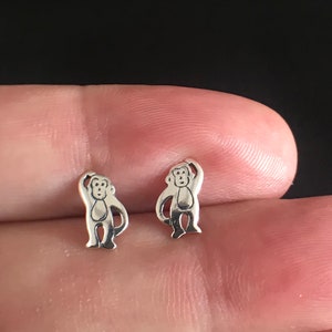 Könnte beinhalten: Ein Paar silberfarbene Ohrstecker mit Affenmotiv. Jeder Ohrring zeigt eine kleine, stilisierte Affenfigur mit erhobenem Arm. Die Ohrringe werden vor einem unscharfen Hintergrund gezeigt.