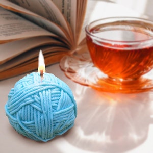 May include: A blue yarn ball candle with a lit wick. The candle is on a white surface with a book and a cup of tea in the background.