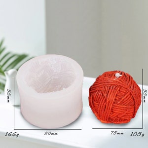 May include: A white silicone mould for making a yarn ball candle. The mould is 5.5 inches tall and 8 inches wide. A red yarn ball candle is shown next to the mould. The candle is 5.5 inches tall and 7.5 inches wide.