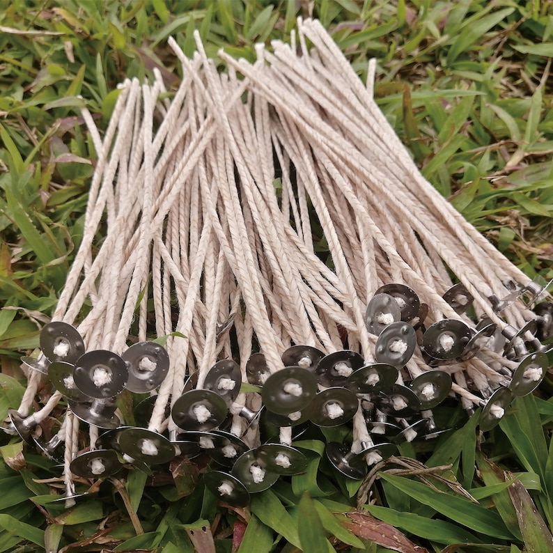 Pode incluir: Um monte de pavios de vela de algod&atilde;o branco com abas de metal presas &agrave;s extremidades. Os pavios est&atilde;o dispostos em uma pilha sobre um leito de grama verde.