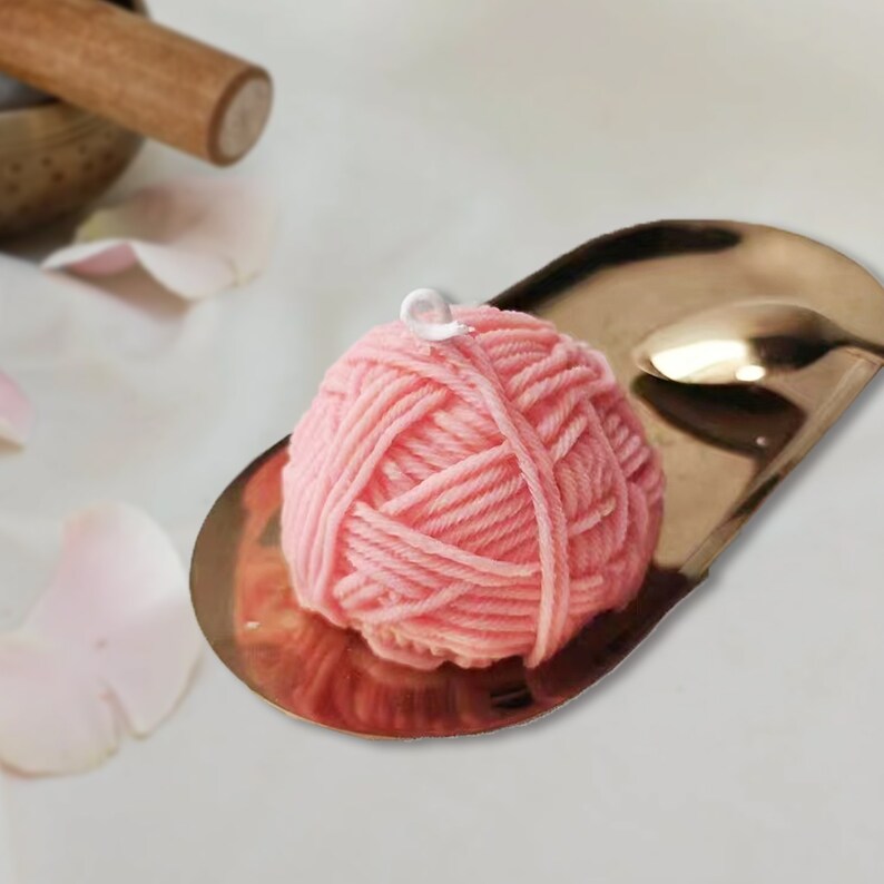May include: A pink ball of yarn with a white loop on top, resting on a gold metal tray.