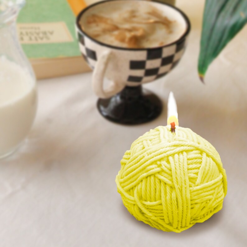 May include: A yellow yarn ball candle with a lit wick. The candle is shaped like a ball of yarn and has a textured surface.
