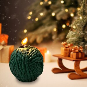 May include: A green and gold yarn ball candle with a lit wick. The candle is sitting on a white surface in front of a blurred background of a Christmas tree and presents.
