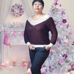 Puede incluir: Una mujer lleva un su&eacute;ter marr&oacute;n con ribetes blancos, pantalones negros y un fondo blanco. El su&eacute;ter tiene cuello en V y un patr&oacute;n texturizado. La mujer est&aacute; de pie frente a un &aacute;rbol de Navidad blanco decorado con adornos rosas y morados.