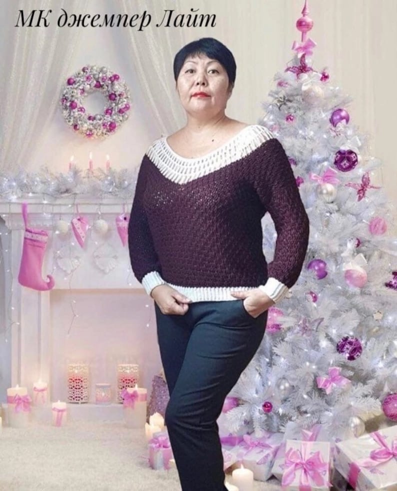 Puede incluir: Una mujer lleva un su&eacute;ter marr&oacute;n con ribetes blancos, pantalones negros y un fondo blanco. El su&eacute;ter tiene cuello en V y un patr&oacute;n texturizado. La mujer est&aacute; de pie frente a un &aacute;rbol de Navidad blanco decorado con adornos rosas y morados.