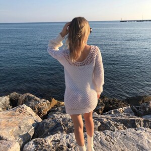 Puede incluir: Una mujer con un vestido de punto blanco y botas blancas se encuentra de pie sobre rocas junto al mar. El agua es azul y el cielo est&aacute; despejado.