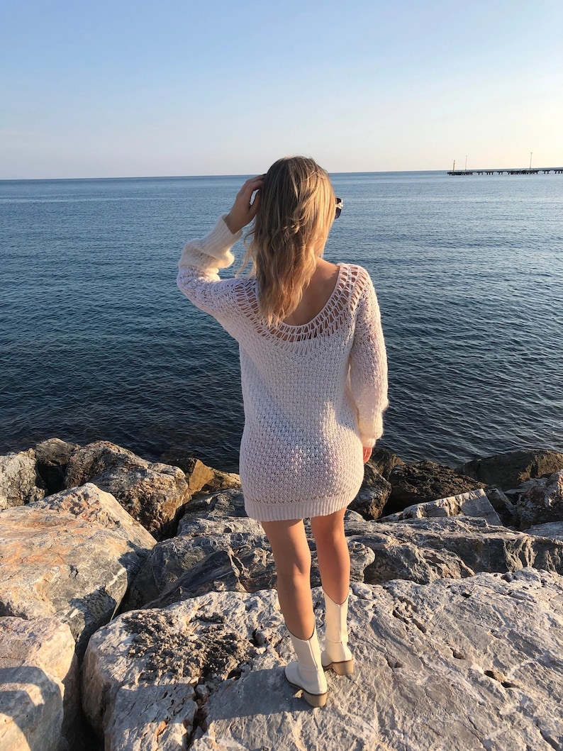 Puede incluir: Una mujer con un vestido de punto blanco y botas blancas se encuentra de pie sobre rocas junto al mar. El agua es azul y el cielo est&aacute; despejado.