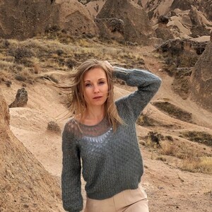 Puede incluir: Una mujer con el pelo largo y rubio que lleva un jersey de punto gris con una superposici&oacute;n de encaje blanco y pantalones beige, de pie en un paisaje rocoso.