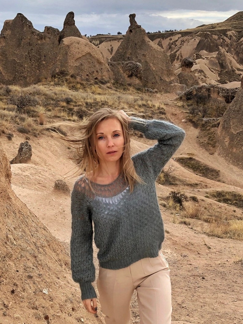 Puede incluir: Una mujer con el pelo largo y rubio que lleva un jersey de punto gris con una superposici&oacute;n de encaje blanco y pantalones beige, de pie en un paisaje rocoso.