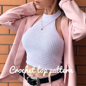 May include: White crochet halter top with a high neckline. The top is cropped and has a loose, open weave. The model is wearing the top with a pink blazer and black belt.