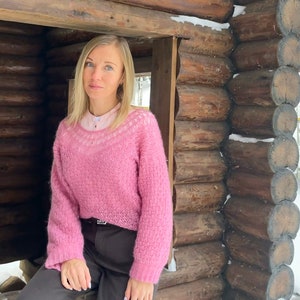 Puede incluir: Una mujer con un su&eacute;ter rosa peludo con ribete de encaje blanco, pantalones marrones y una camisa blanca debajo. Est&aacute; sentada en una caba&ntilde;a de madera con nieve en los troncos.