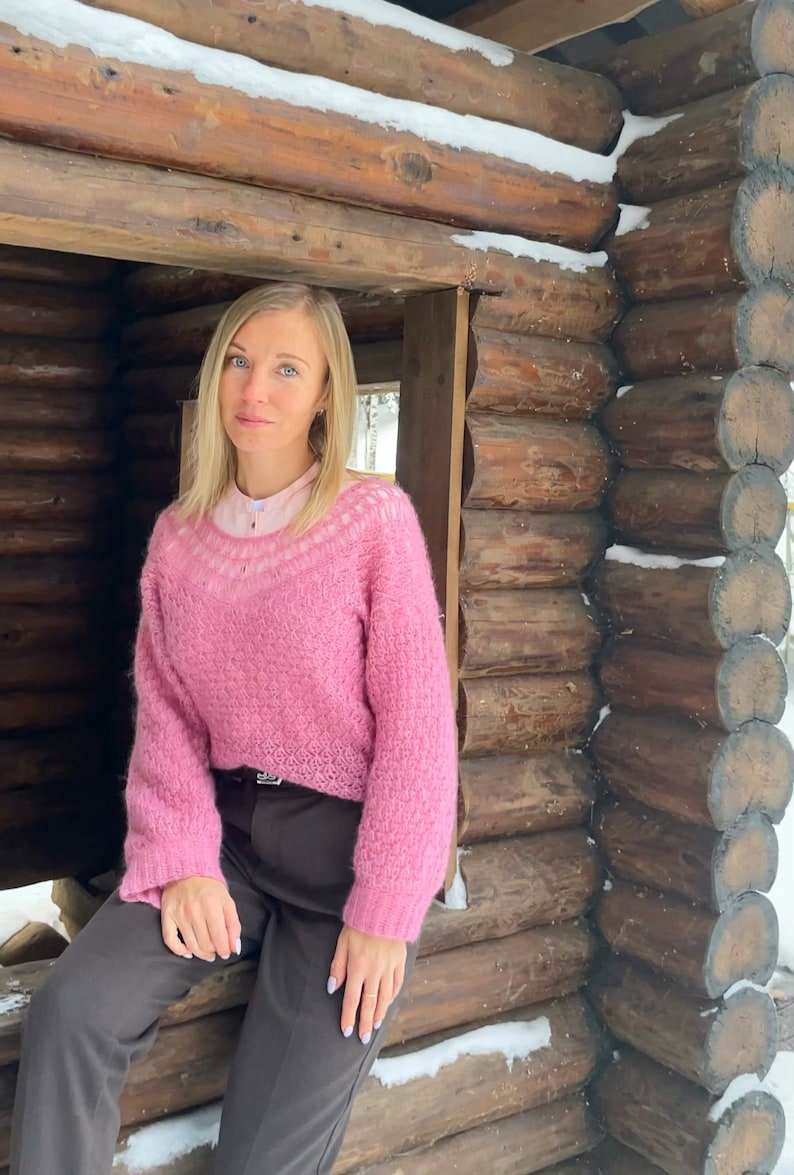 Puede incluir: Una mujer con un su&eacute;ter rosa peludo con ribete de encaje blanco, pantalones marrones y una camisa blanca debajo. Est&aacute; sentada en una caba&ntilde;a de madera con nieve en los troncos.