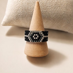 Puede incluir: Un anillo elaborado con pequeñas cuentas cuadradas en un patrón repetitivo. El diseño presenta un motivo de diamante central creado con cuentas de color plata metálica sobre un fondo de cuentas negras mate, formando una banda ancha y plana.