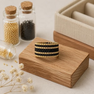 Puede incluir: Un anillo ancho elaborado con pequeñas cuentas cilíndricas dispuestas en franjas horizontales alternas de color oro metálico y negro mate. El anillo presenta una textura uniforme similar a un tejido gracias a la alineación de las cuentas.