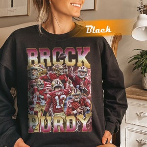 May include: Black sweatshirt featuring a collage of football players in red and gold uniforms. The sweatshirt has the name "BROCK PURDY" in a vintage-style font. The word "Black" is written in orange on the top right.