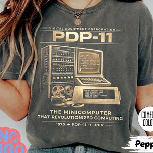 May include: A dark grey t-shirt featuring a vintage computer graphic with the text "PDP-11". The design includes a computer, monitor, and text that reads "THE MINICOMPUTER THAT REVOLUTIONIZED COMPUTING". The shirt is labeled "Comfort Colors" and "Pepper".